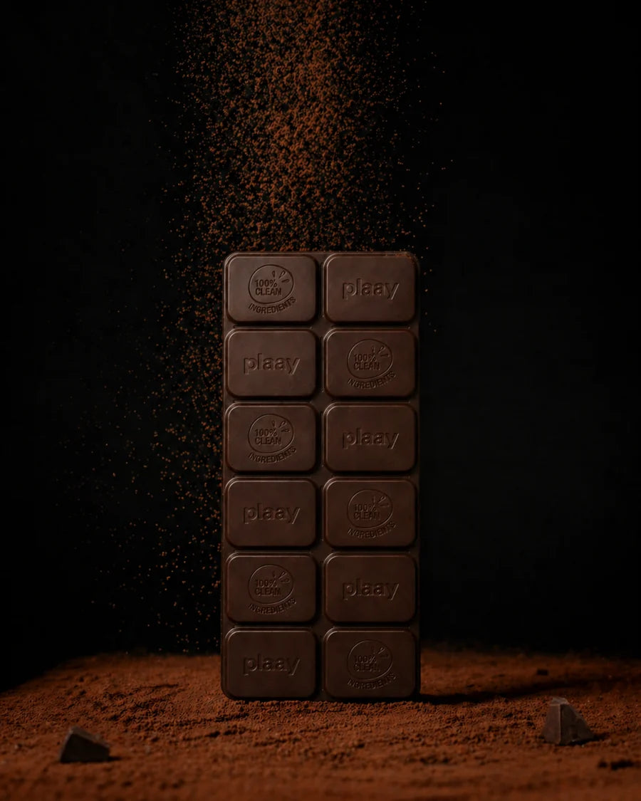 A vertical stack of dark chocolate squares with the brand name 'plaay' embossed on each piece, standing upright on a bed of cocoa powder against a black background with cocoa powder falling above.