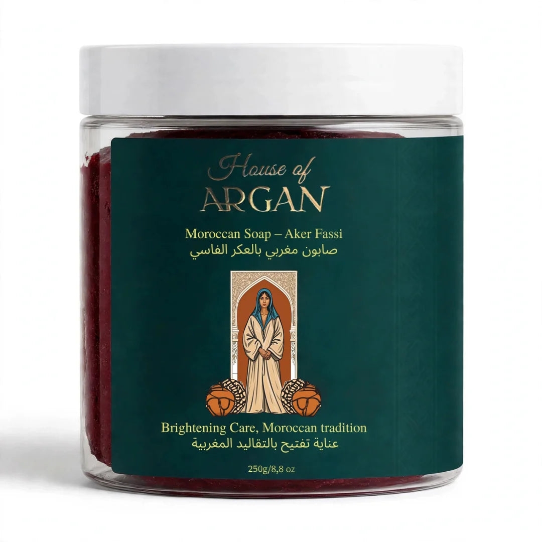 A clear plastic jar of House of Argan Moroccan Soap (Aker Fassi) with a white lid and a dark green label featuring gold lettering and a central illustration, set against a plain white background.