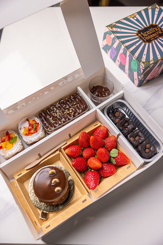An open white bakery gift box filled with assorted desserts, including a chocolate cake with a bear face, strawberries, chocolate brownies, and small fruit tart cups. A branded outer box sits beside the open box on a light marble surface.