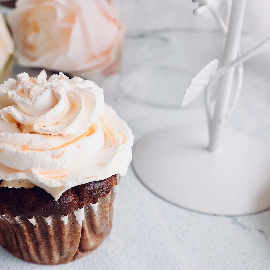 A close-up of a frosted cupcake with white and pale orange icing in a brown paper liner, set on a light marble-like surface with a soft-focus white decorative object in the background.