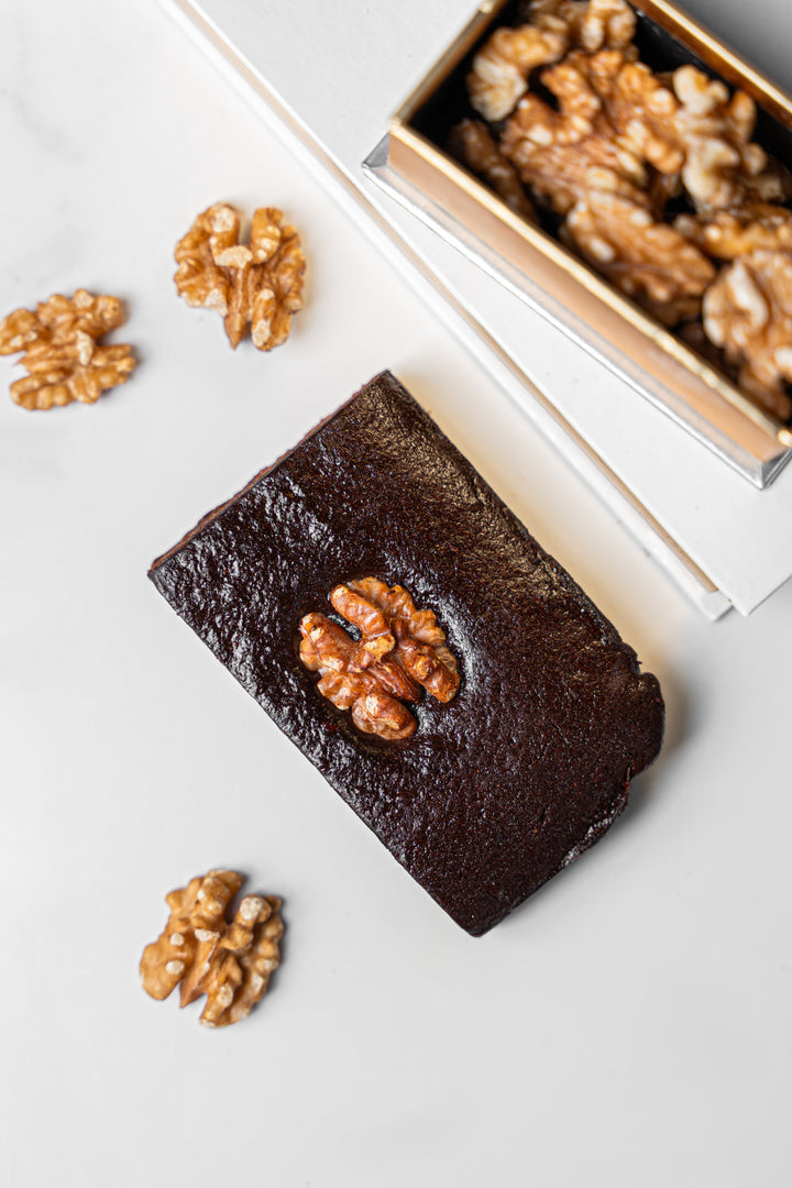 A square dark chocolate brownie topped with a whole walnut, arranged on a light marble surface next to an open box of walnuts.
