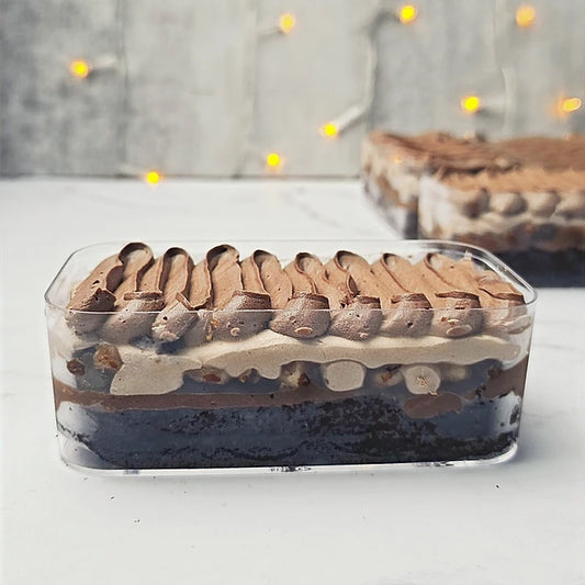 A rectangular clear plastic container holding a layered chocolate dessert with dark chocolate cake, cream-colored filling, and piped chocolate swirls on top, set on a light surface with a blurred cake and warm string lights in the background.