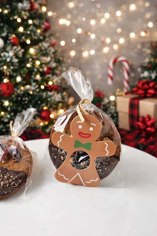 A Christmas-themed cookie gift bag featuring a gingerbread man cutout with a green bow tie, filled with chocolate chip cookies and wrapped in clear cellophane, set against a festive holiday background with a decorated tree and lights.
