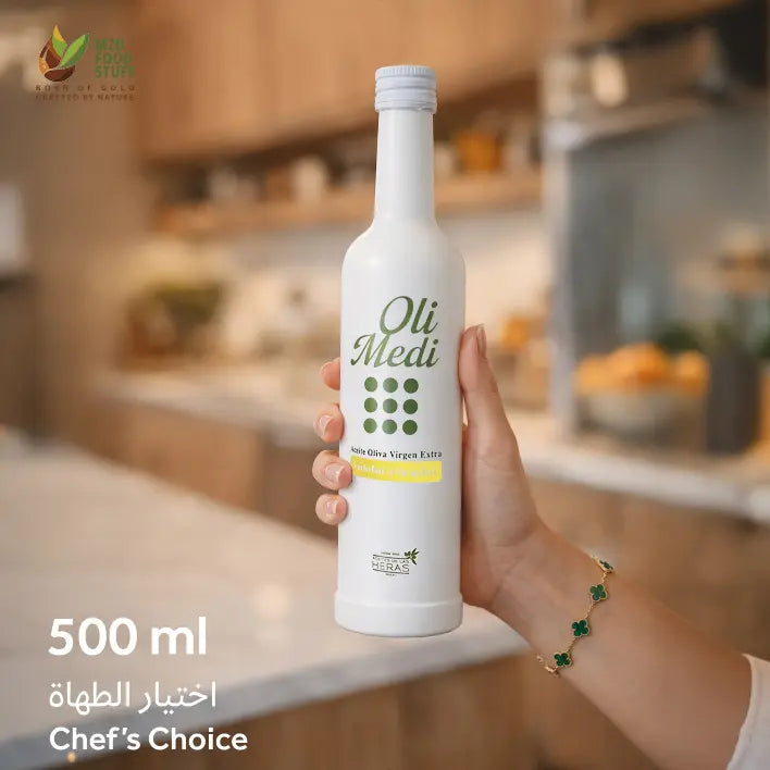 A hand holds a 500 ml bottle of Oli Medi non-alcoholic virgin olive oil in a bright, modern kitchen setting. The bottle is white with green branding text and a yellow accent label.