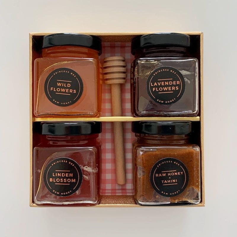 A boxed honey gift set containing four glass jars of honey (Wild Flowers, Lavender Flowers, Linden Blossom, and Raw Honey Tahini) with a wooden honey dipper and a wooden divider, presented in a light brown box on a white background.