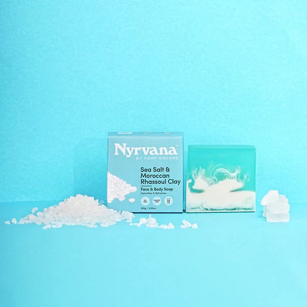 Nyrvana Sea Salt & Moroccan Rhassoul Clay face & body soap shown with its packaging box and a bar of soap on a bright blue background, with white soap crystals scattered in front.