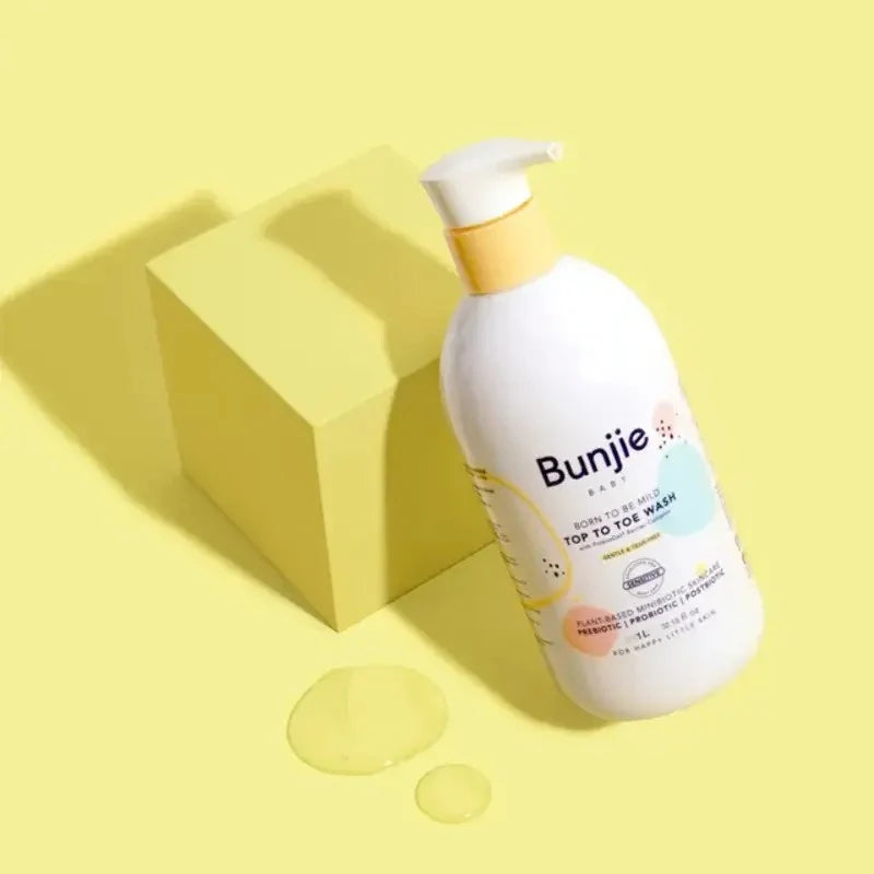 A bottle of Bunjie Baby Top 10 Ice Wash hand sanitizer with a white pump dispenser and yellow cap, styled on a yellow background with a yellow block and liquid droplets.