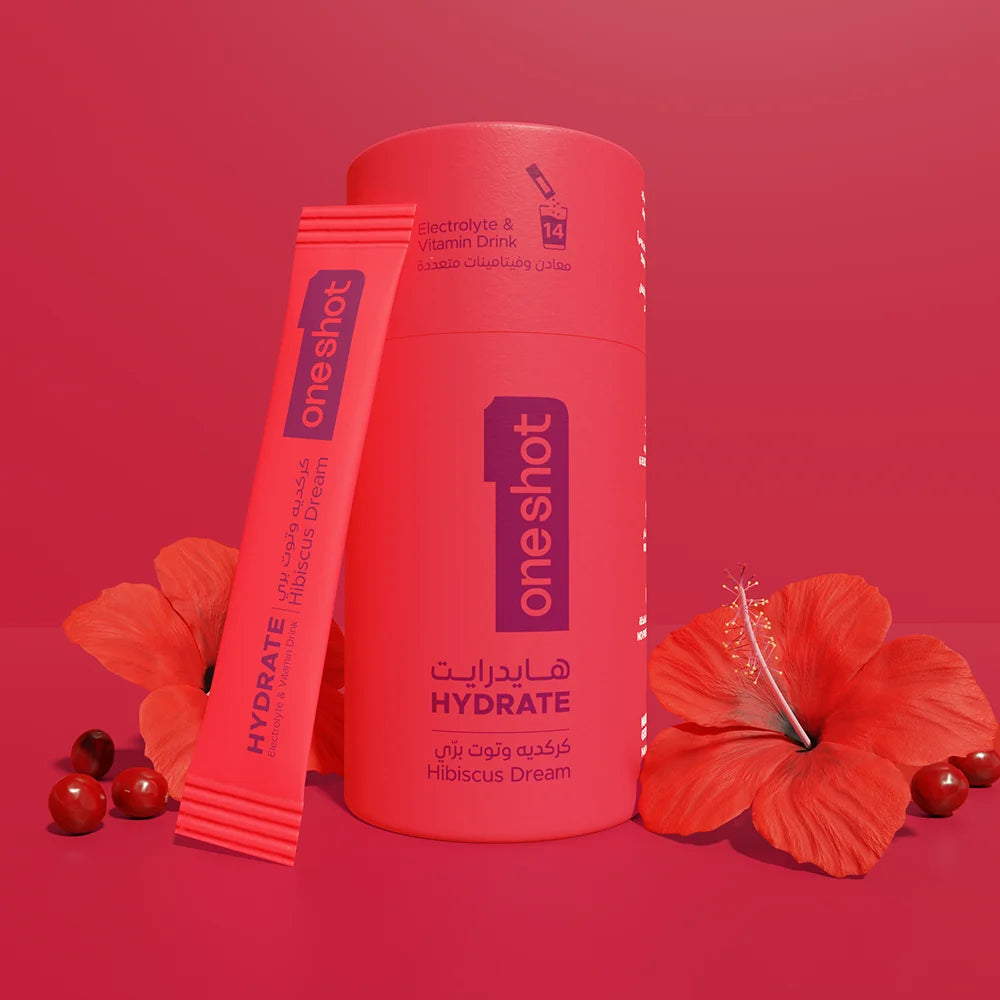 A cylindrical bottle and a single stick pack of one shot Hydrate electrolyte drink mix, both in a bright coral-pink color, styled with red hibiscus flowers and small red berries on a matching coral-pink background.