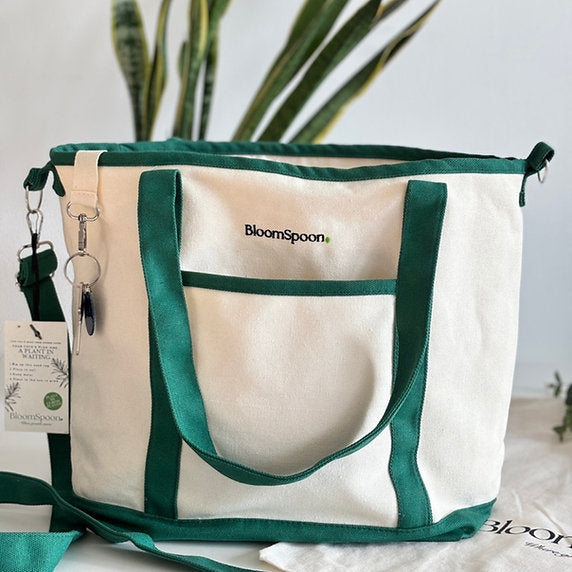 A cream canvas tote bag with dark green trim, handles, and pocket borders. The bag features the 'BloomSpoon' brand name printed in black on the front pocket.