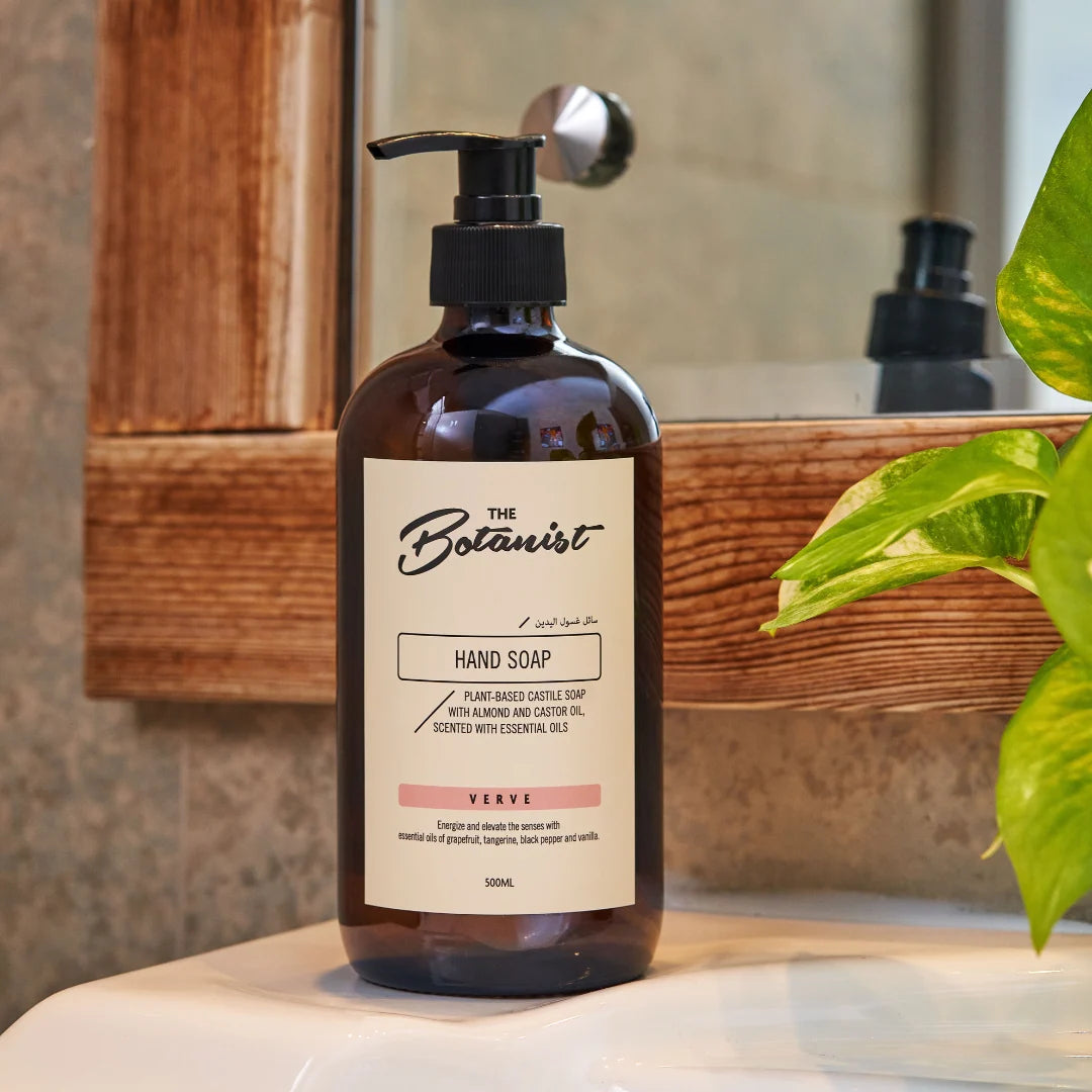A 300ml pump bottle of 'The Botanist' hand soap in the 'Sage' scent, placed on a white bathroom counter in front of a wooden mirror with a green plant leaf in the foreground.