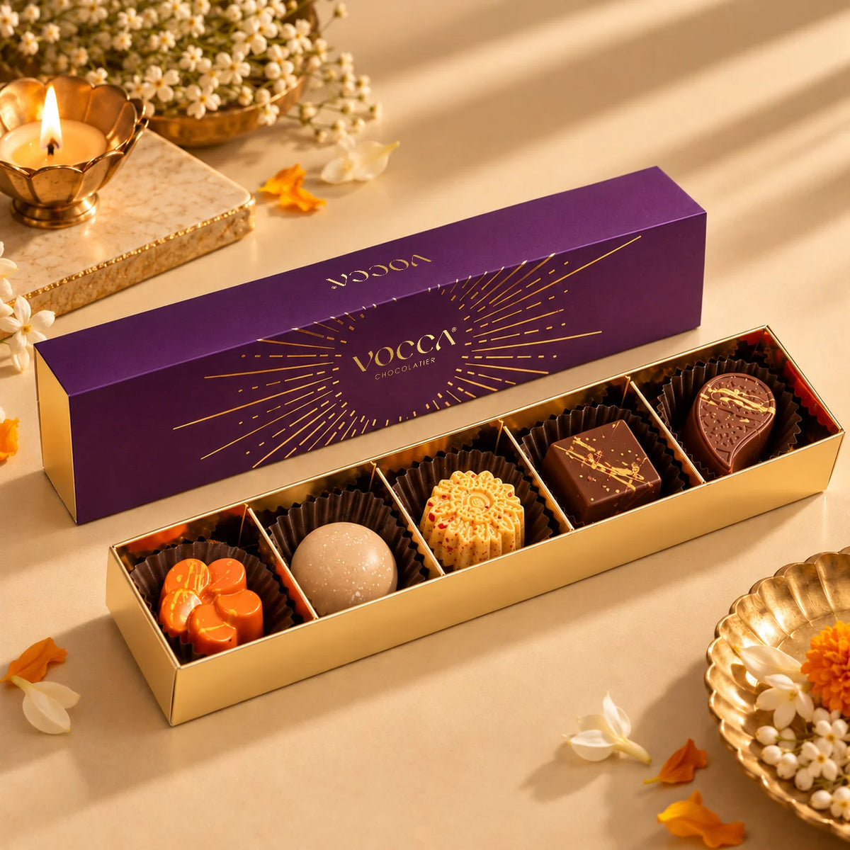 A luxury chocolate gift set by VOGTA, featuring a purple and gold branded box alongside an open tray of assorted gourmet chocolates, styled with warm decorative props on a beige surface.