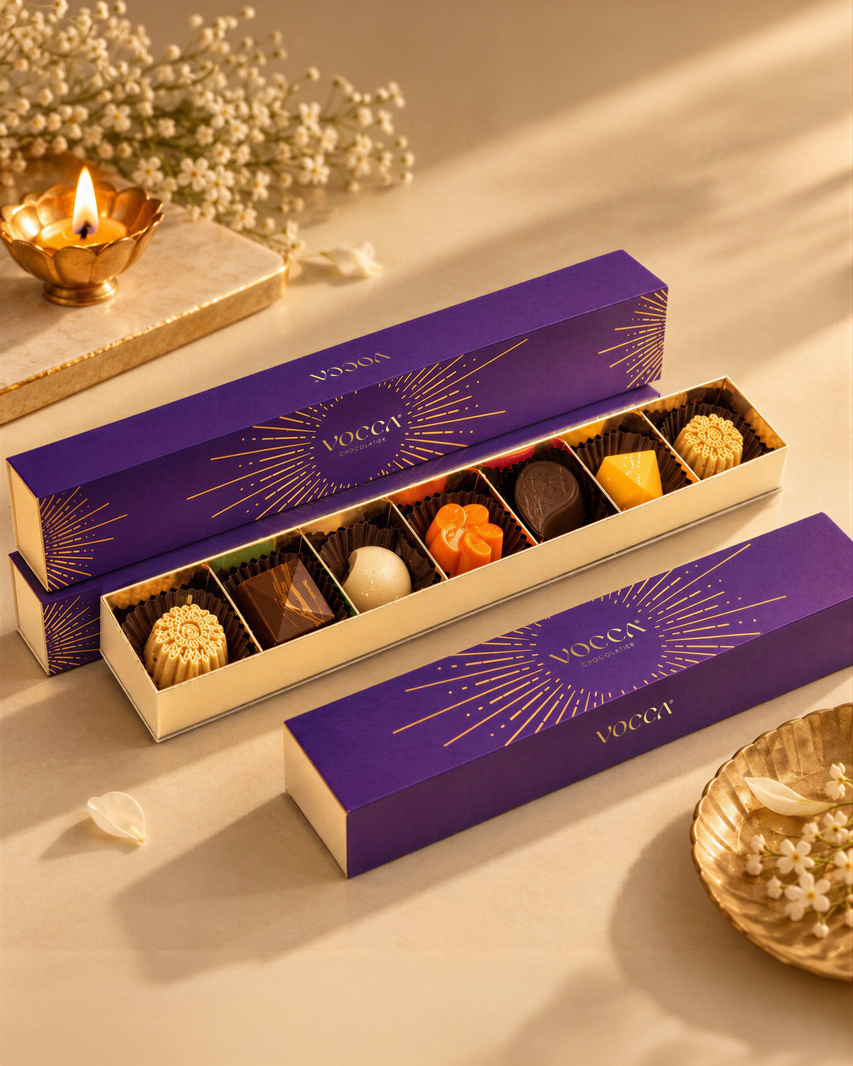 Two purple and gold boxes of chocolates arranged on a warm beige surface with decorative dried flowers and a small gold candle. One box is open, showing assorted truffles and chocolates in paper cups.