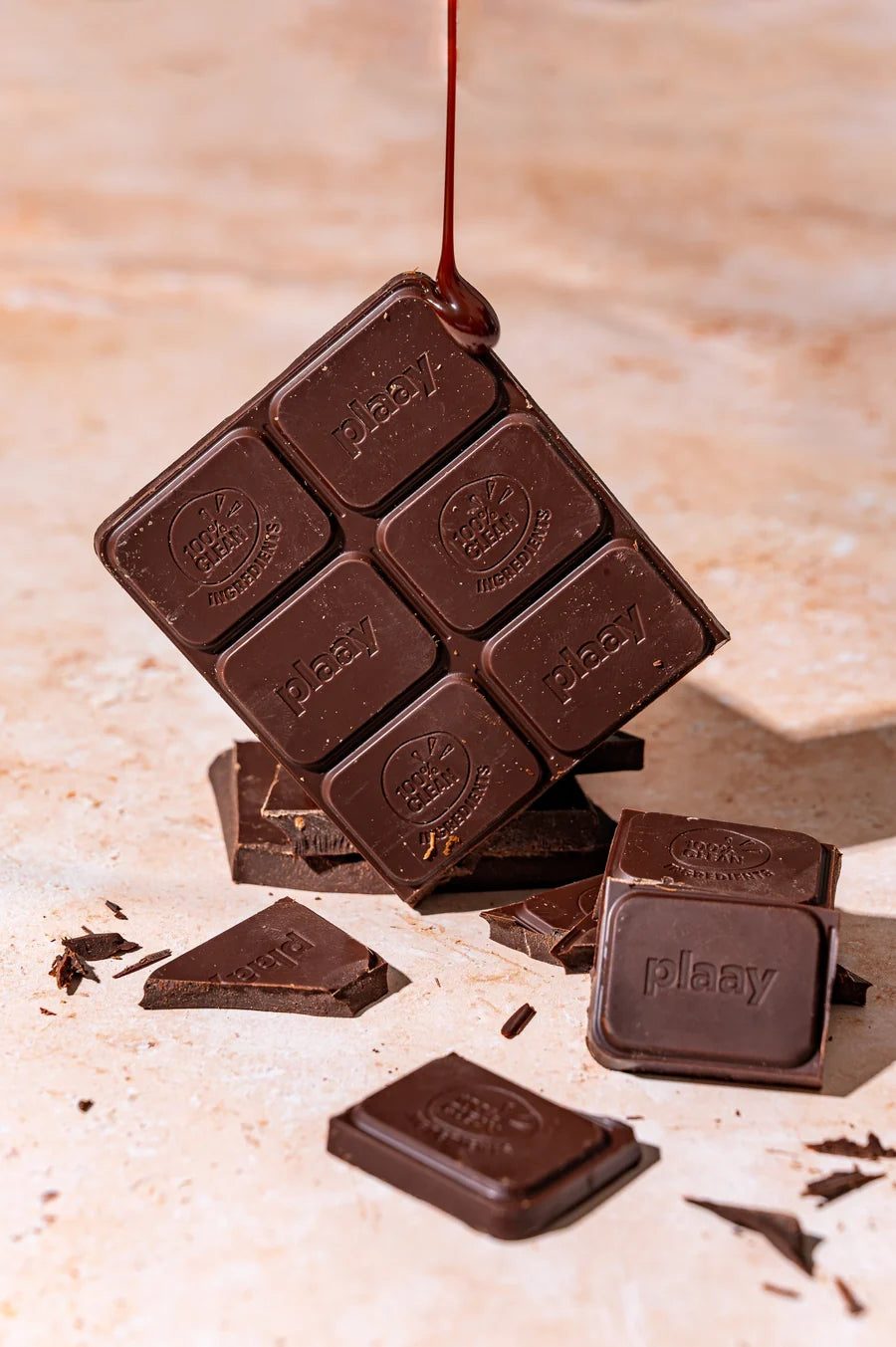 A square dark chocolate bar with the brand name 'plaay' embossed on each piece, shown tilted with chocolate syrup dripping from the top, alongside scattered broken chocolate pieces on a light beige stone surface.