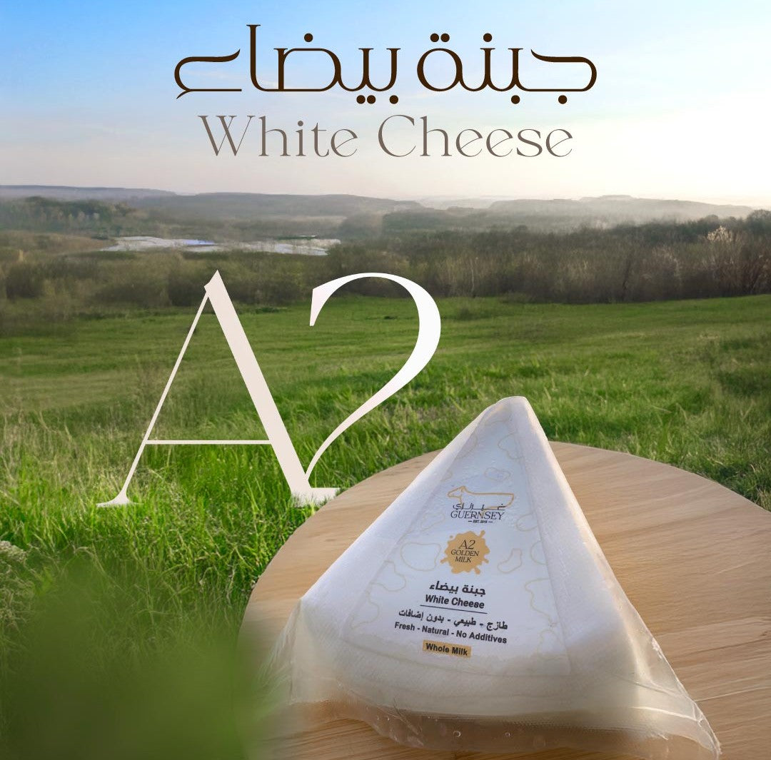 A triangular wedge of white cheese wrapped in clear plastic sits on a round wooden board in front of a blurred outdoor landscape with green fields and hills. Large overlaid text reads 'White Cheese' and 'A2', with Arabic text at the top.