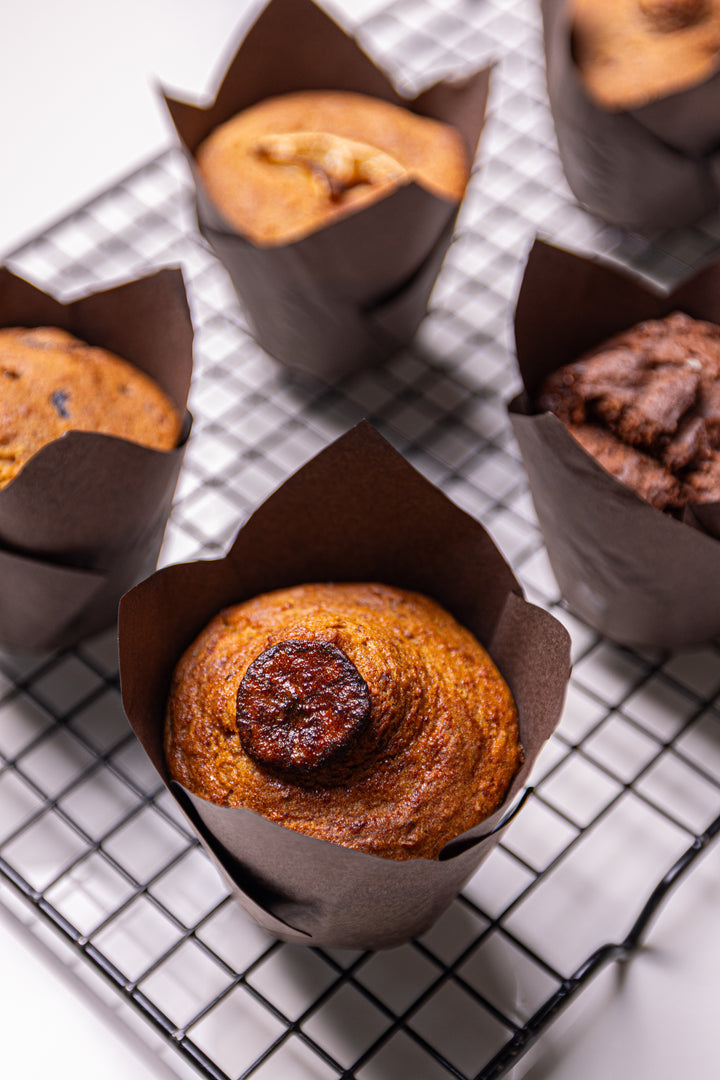 Freshly baked pumpkin spice muffins in brown paper liners, with one in the foreground on a black wire cooling rack and more muffins blurred in the background.