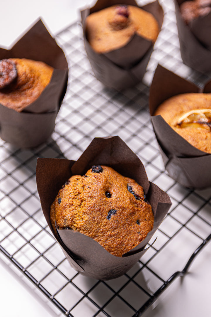 Several golden-brown muffins in dark brown paper liners rest on a black wire cooling rack over a white surface.