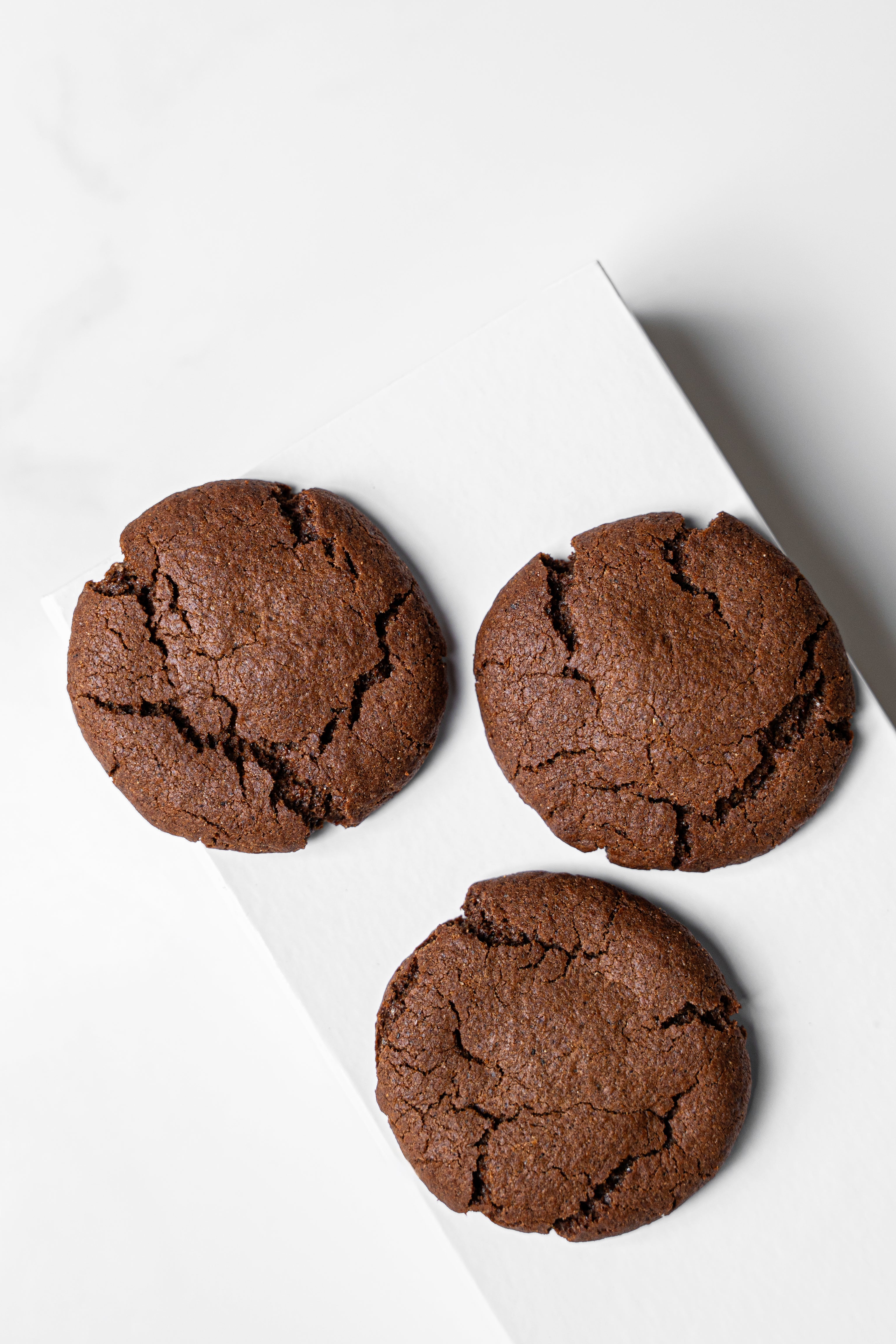 Three chocolate chip cookies with a cracked, baked surface arranged on a white diagonal board against a light gray background.