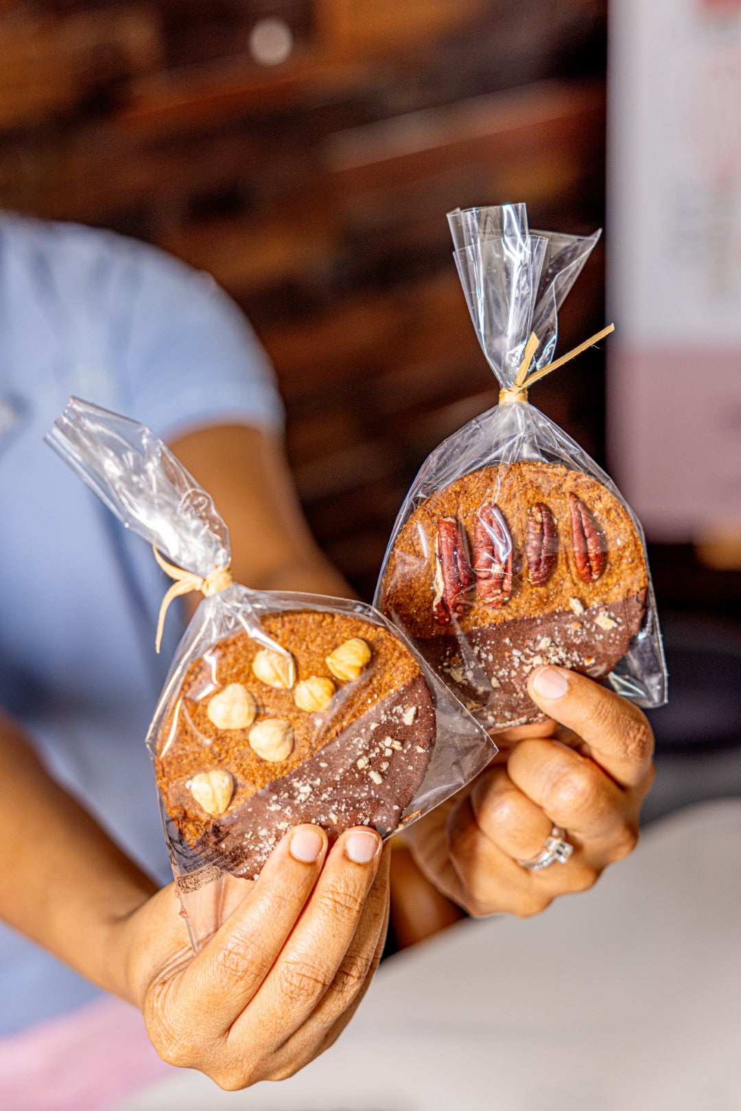 Two small artisanal cookies wrapped in clear cellophane bags tied with wooden sticks, held by hands against a blurred indoor background.