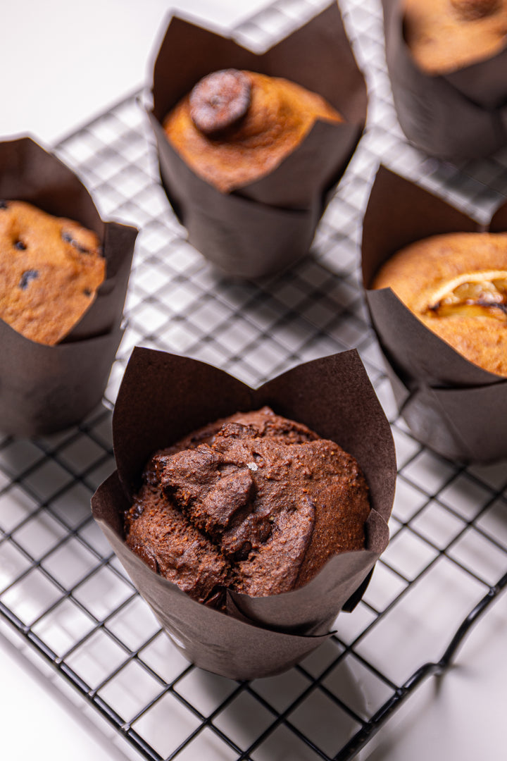 Several chocolate chip muffins in dark brown paper liners, with one in the foreground on a black wire cooling rack over a white surface.