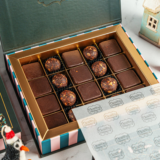 An open box of assorted chocolates on a light marble surface. The box has a dark green lid, a gold inner tray, and teal, white, and pink striped sides.
