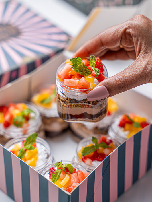 A hand with pink manicured nails holds a small glass jar of layered dessert topped with fresh fruit and mint, with several similar dessert jars inside a pink and black striped box.