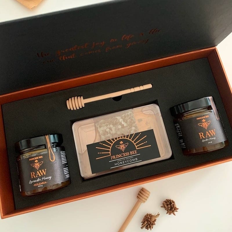 A honey gift set in an open black presentation box with a copper-toned inner tray. The set includes two dark glass jars of honey with black lids, a clear plastic container of honeycomb, a wooden honey dipper, and two small pinecones on a white surface.