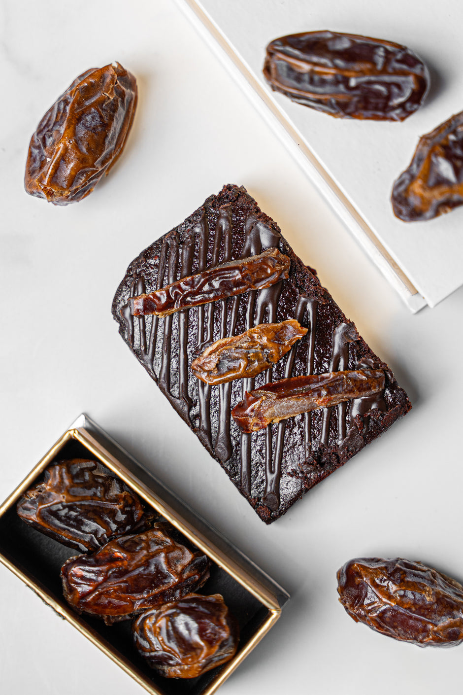 A styled flat-lay of chocolate brownies topped with whole dates, alongside loose dates and open gift boxes on a light marble surface.