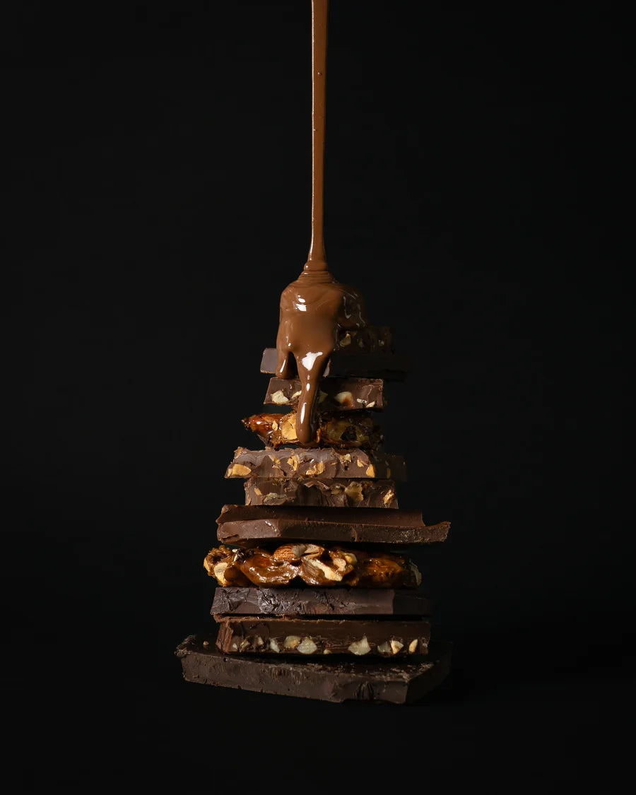 A vertical stack of assorted chocolate bars and nuts, with glossy caramel or milk chocolate drizzled down the center, set against a solid black background.