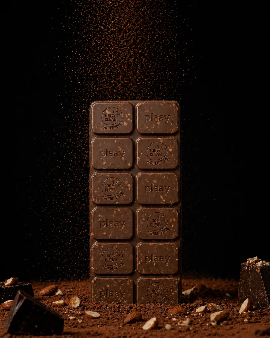 A vertical bar of dark chocolate with the brand name 'plaav' embossed on each square, standing upright on a dark surface with cocoa powder and chocolate pieces scattered around it.
