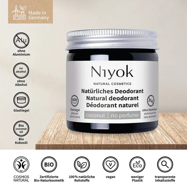 A jar of Niyok Natural Cosmetics coconut deodorant is shown on a wooden surface with a beige background, surrounded by German certification and feature icons.