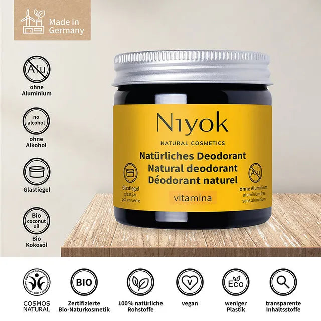 A jar of Niyok Natural Cosmetics vitamina natural deodorant, featuring a yellow label and silver cap, displayed on a wooden surface with German certification icons on a beige background.