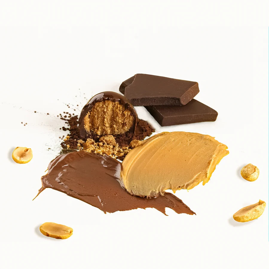 A styled food product shot of a chocolate-covered peanut butter ball, broken to show the peanut butter filling, with chocolate chunks, peanut butter spread, and scattered peanuts on a white background.