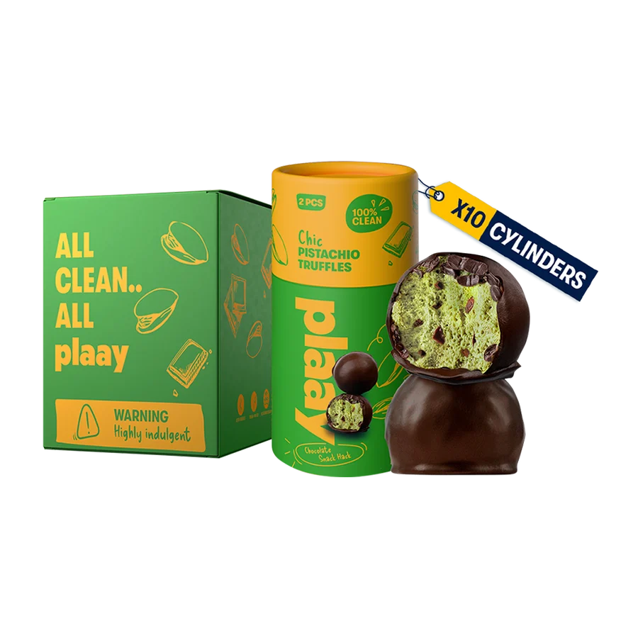 A product display of PlaaY Pistachio Truffle candies, featuring a green and yellow cardboard box, a matching green and yellow cylindrical tin, and two chocolate-covered pistachio truffles (one whole and one split open to show the green pistachio filling).