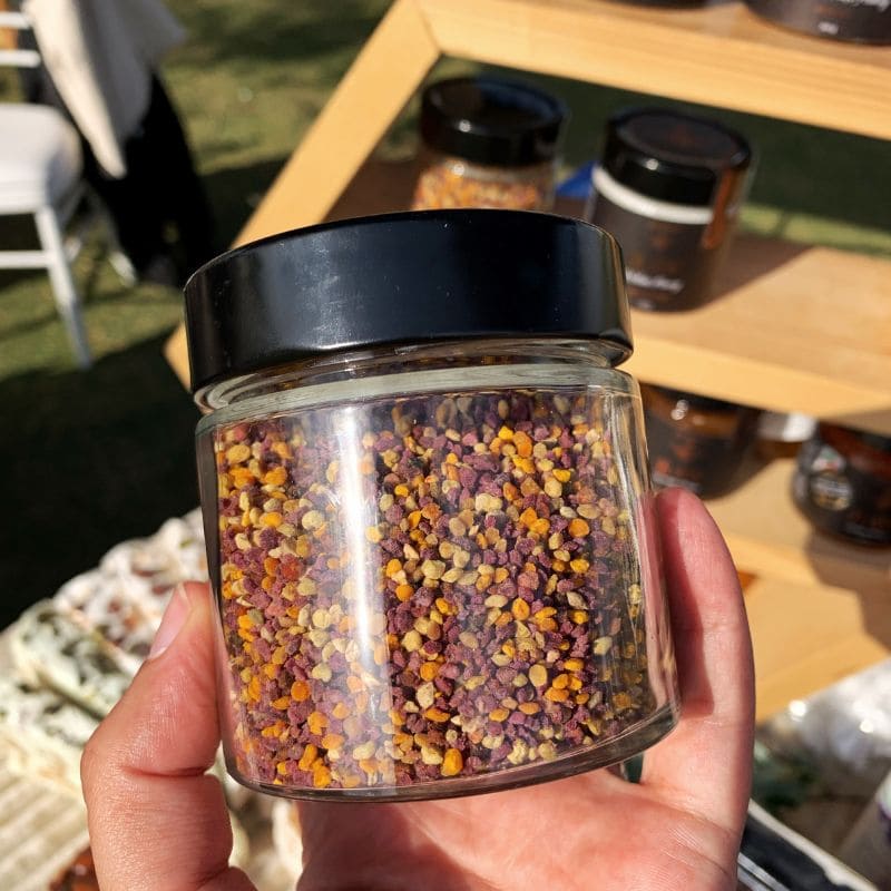 A hand holding a clear glass jar filled with multicolored bee pollen granules, topped with a black lid, set against an outdoor market background with wooden tables and other jars.