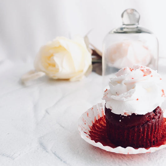 A red velvet cupcake with white frosting sits in a white paper liner on a light surface, with a blurred white rose and a glass jar in the background.