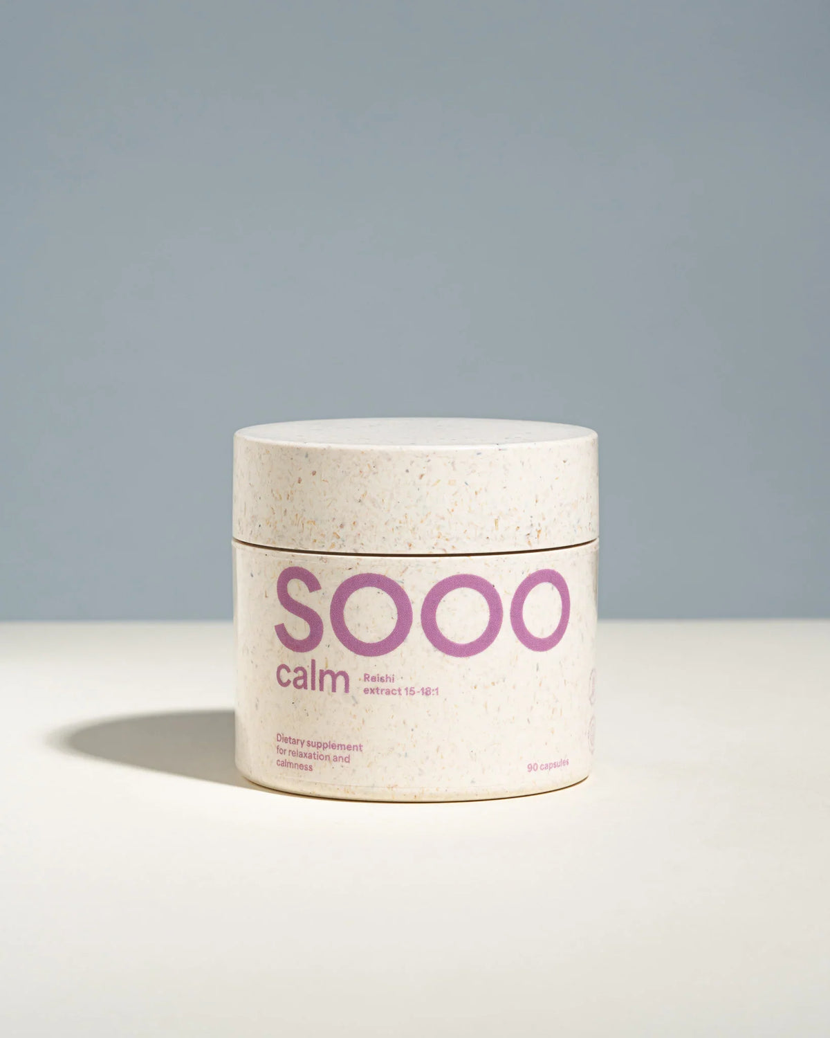 A cylindrical supplement jar labeled 'SOOO calm' in purple text, with a speckled off-white lid and base, set against a clean two-tone gray and white background.