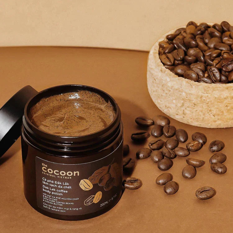 A jar of Cocoon Natural Visions coffee body polish with its lid off, placed on a brown surface next to scattered coffee beans and a bowl of coffee beans.
