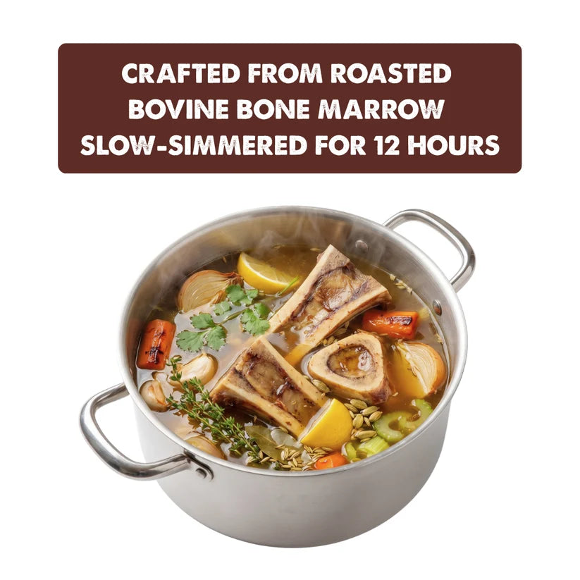 A stainless steel pot filled with a bone marrow soup or stew, featuring visible bone pieces, lemon slices, carrots, herbs, and a brown text banner at the top that reads, 'CRAFTED FROM ROASTED BOVINE BONE MARROW SLOW-SIMMERED FOR 12 HOURS.'.