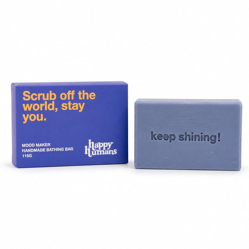 A blue rectangular box with yellow text next to a dusty blue handmade bath bar that reads 'keep shining!'. The box says 'Scrub off the world, stay you.