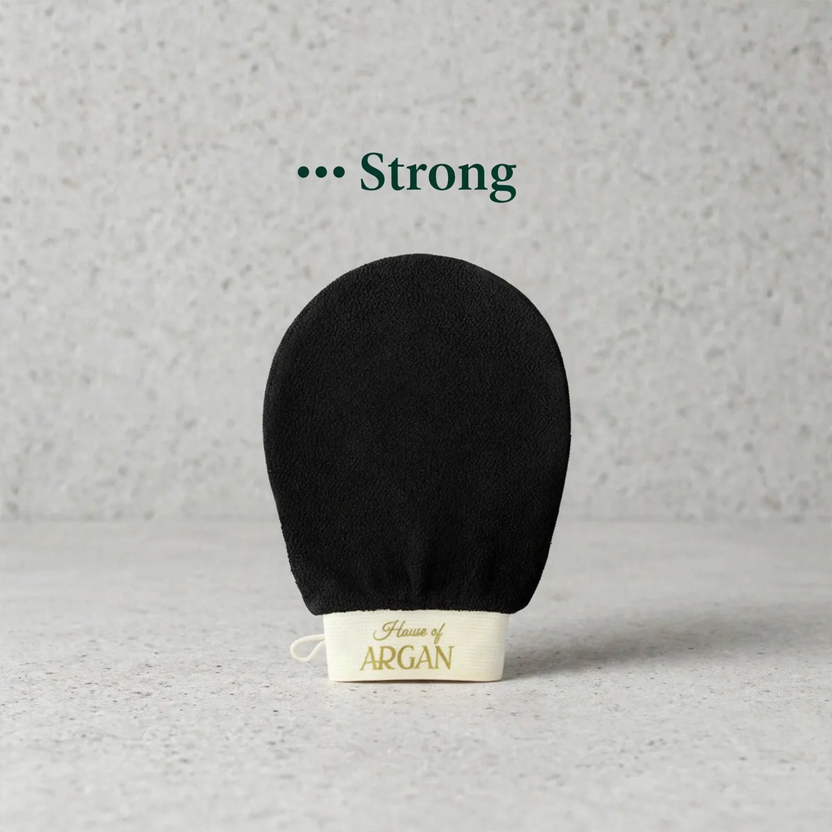A black exfoliating glove with a cream-colored cuff printed with 'House of ARGAN' in gold, set against a textured light gray background with the text '.