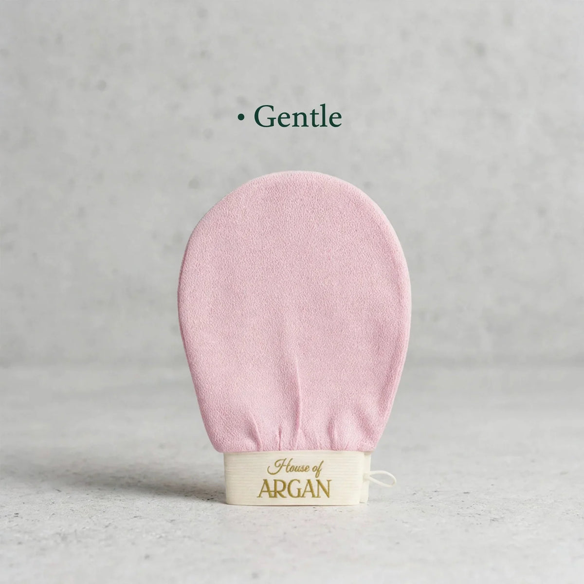 A pale pink exfoliating loofah with a cream-colored fabric band at the base printed with 'House of ARGAN' in gold lettering. The word 'Gentle' is written in dark green text above the product.