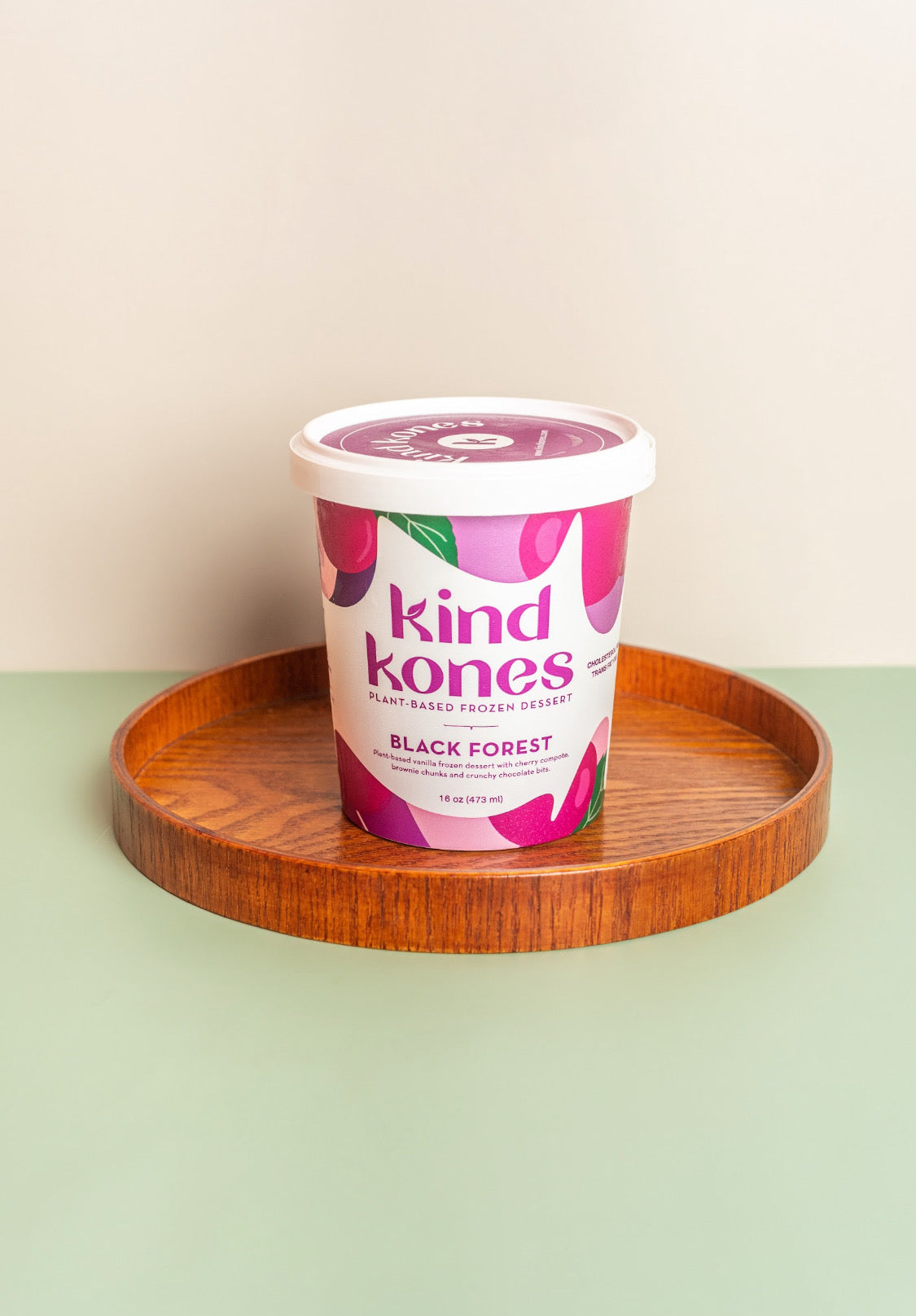 A tub of Kind Kones Black Forest flavored frozen yogurt sits on a round wooden tray against a two-tone beige and sage green background.