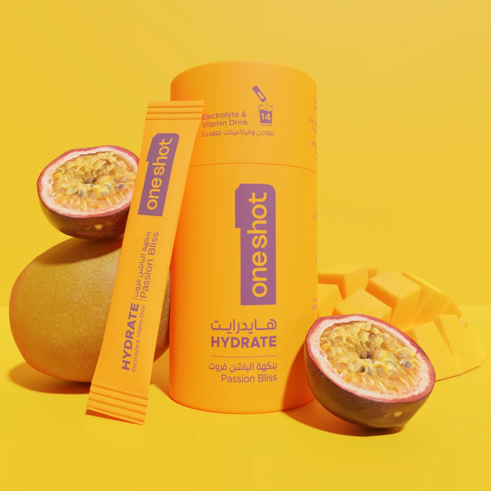 An orange cylindrical bottle and matching stick pack of OneShot Hydrate Passion Bliss drink mix, styled with whole and sliced passion fruits and mango pieces on a bright yellow background.