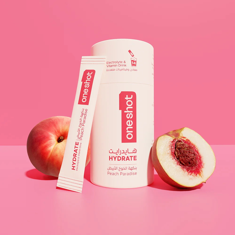 A white cylindrical bottle and matching sachet for One Shot Hydrate Peach Paradise electrolyte drink mix, styled with a whole peach and a halved peach on a solid pink background.