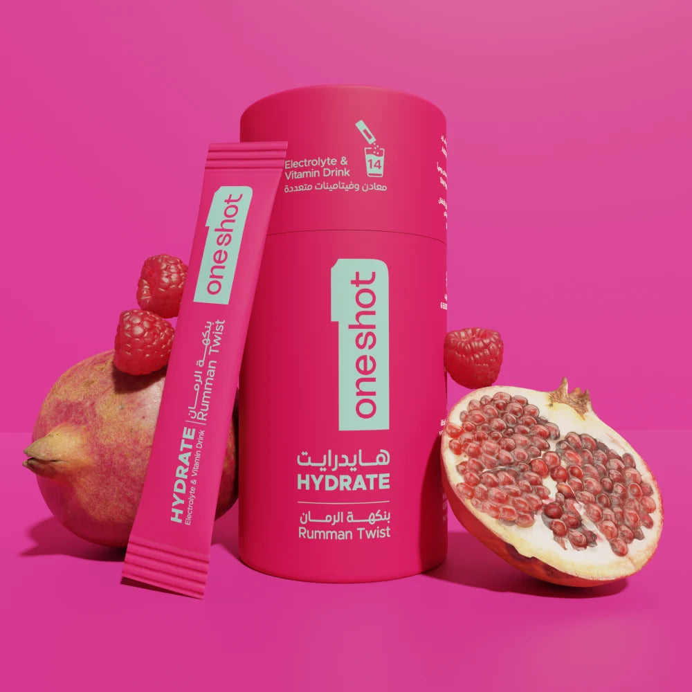 A pink cylindrical bottle and matching sachet of OneShot Hydrate Rumman Twist electrolyte drink mix, styled with whole and halved pomegranates on a solid pink background.