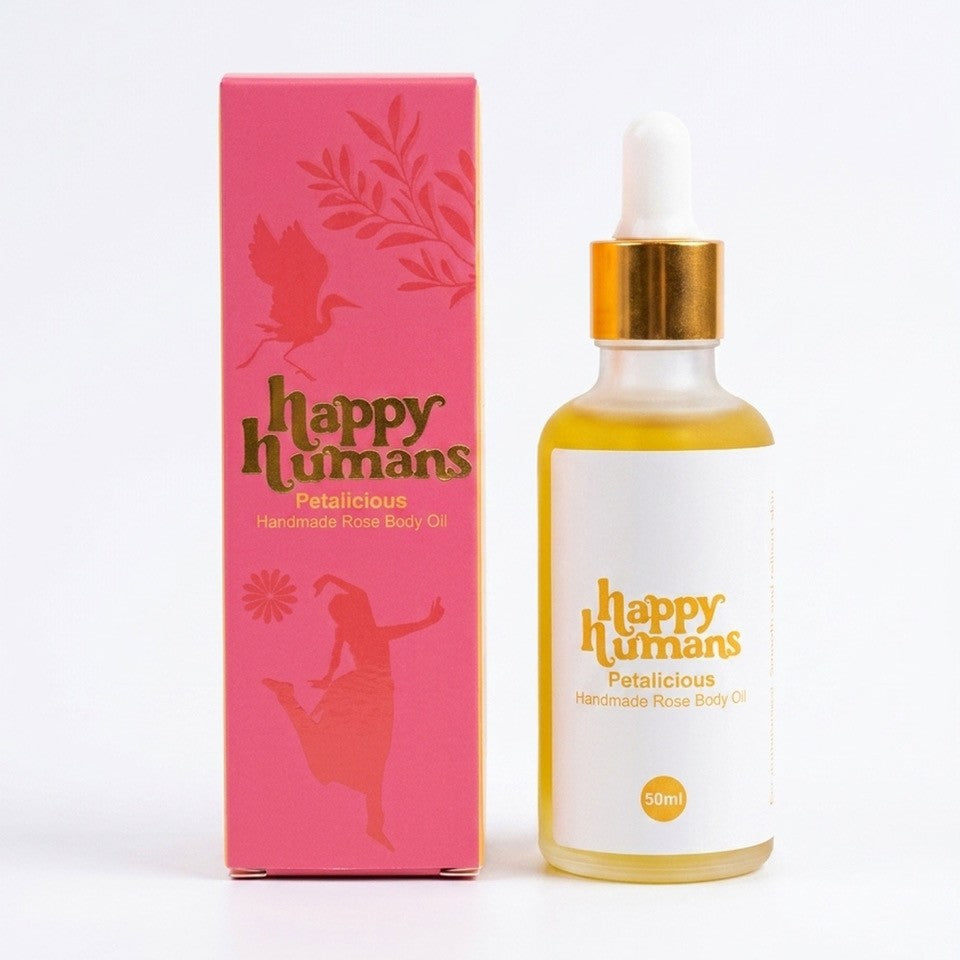 A 50ml bottle of Happy Humans Petalicious Handmade Rose Body Oil next to its pink packaging box, featuring a white dropper cap and gold collar on a white background.