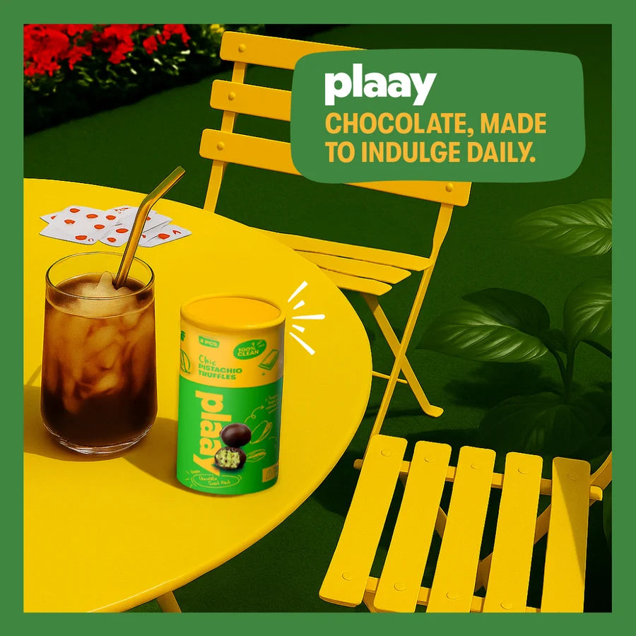 A can of Plaay chocolate on a bright yellow table next to an iced coffee drink, with a yellow chair and a green promotional badge that reads 'plaay CHOCOLATE, MADE TO INDULGE DAILY.'.