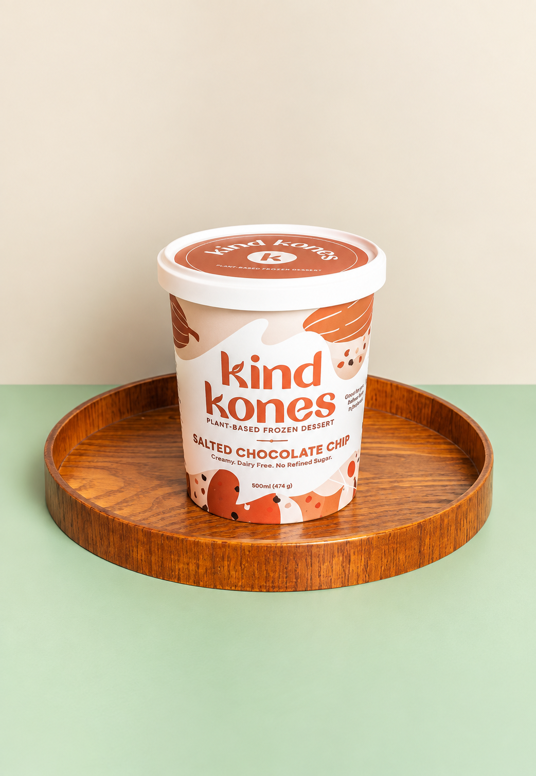 A tub of Kind Kones Salted Chocolate Chip plant-based frozen dessert sits on a round wooden tray against a two-tone beige and sage green background.