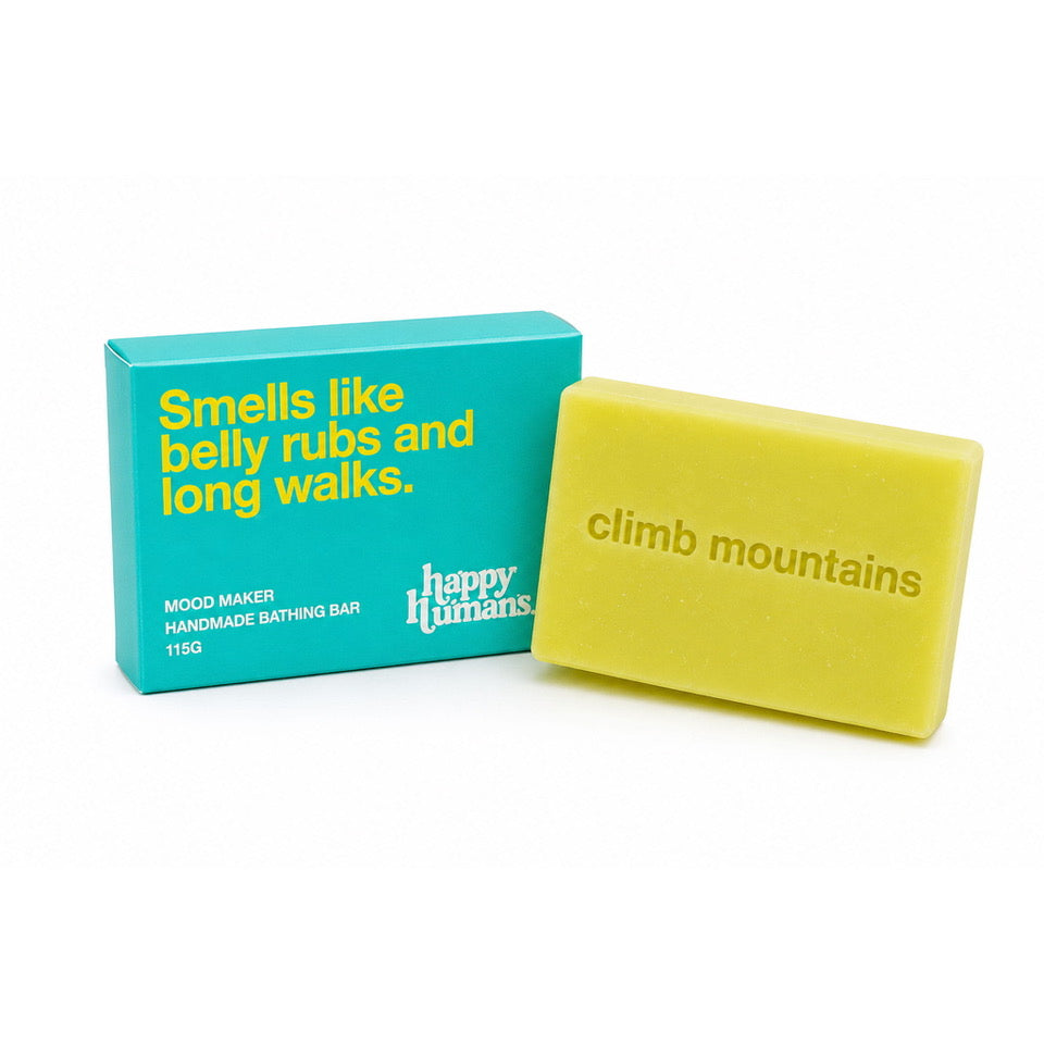 A yellow handmade bath bar next to its turquoise packaging box. The bar is embossed with the brand name 'climb mountains' and the box features yellow text that reads 'Smells like belly rubs and long walks.