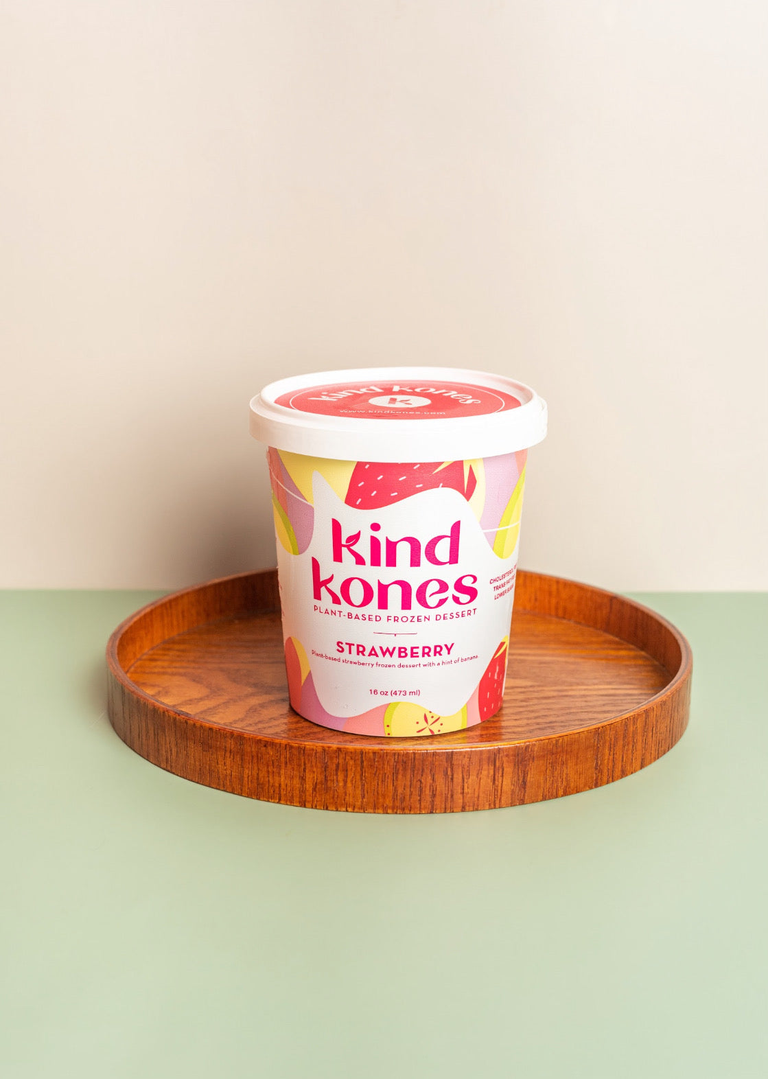 A tub of Kind Kones Strawberry sugar-free protein oats sits on a round wooden tray against a split beige and sage green background.